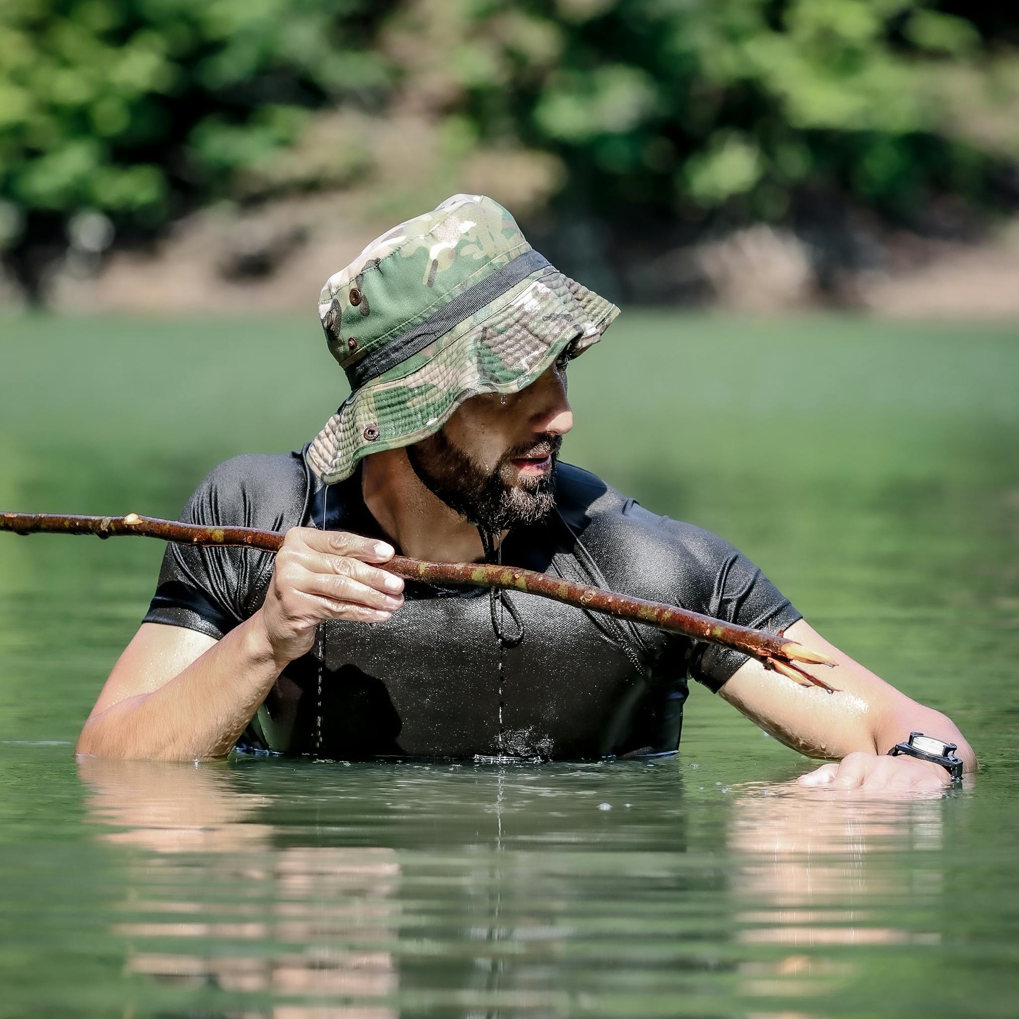 An adventurous man in a camo hat stands in water holding a sharpened stick, showcasing survival skills.