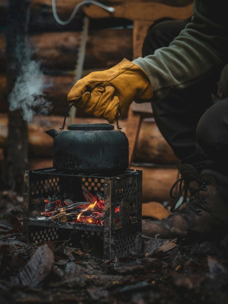 Close-up of a kettle on a camping stove with glove-covered hand adjusting it outdoors.