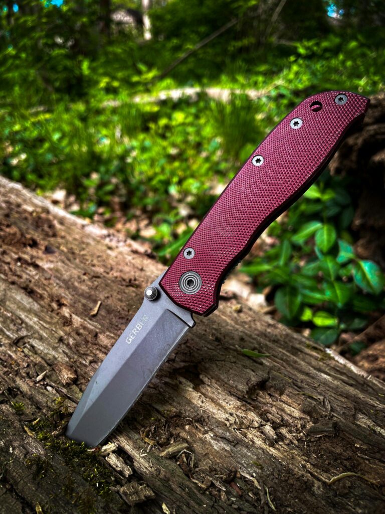Detailed view of a red handled knife embedded in a log in a lush outdoor setting.