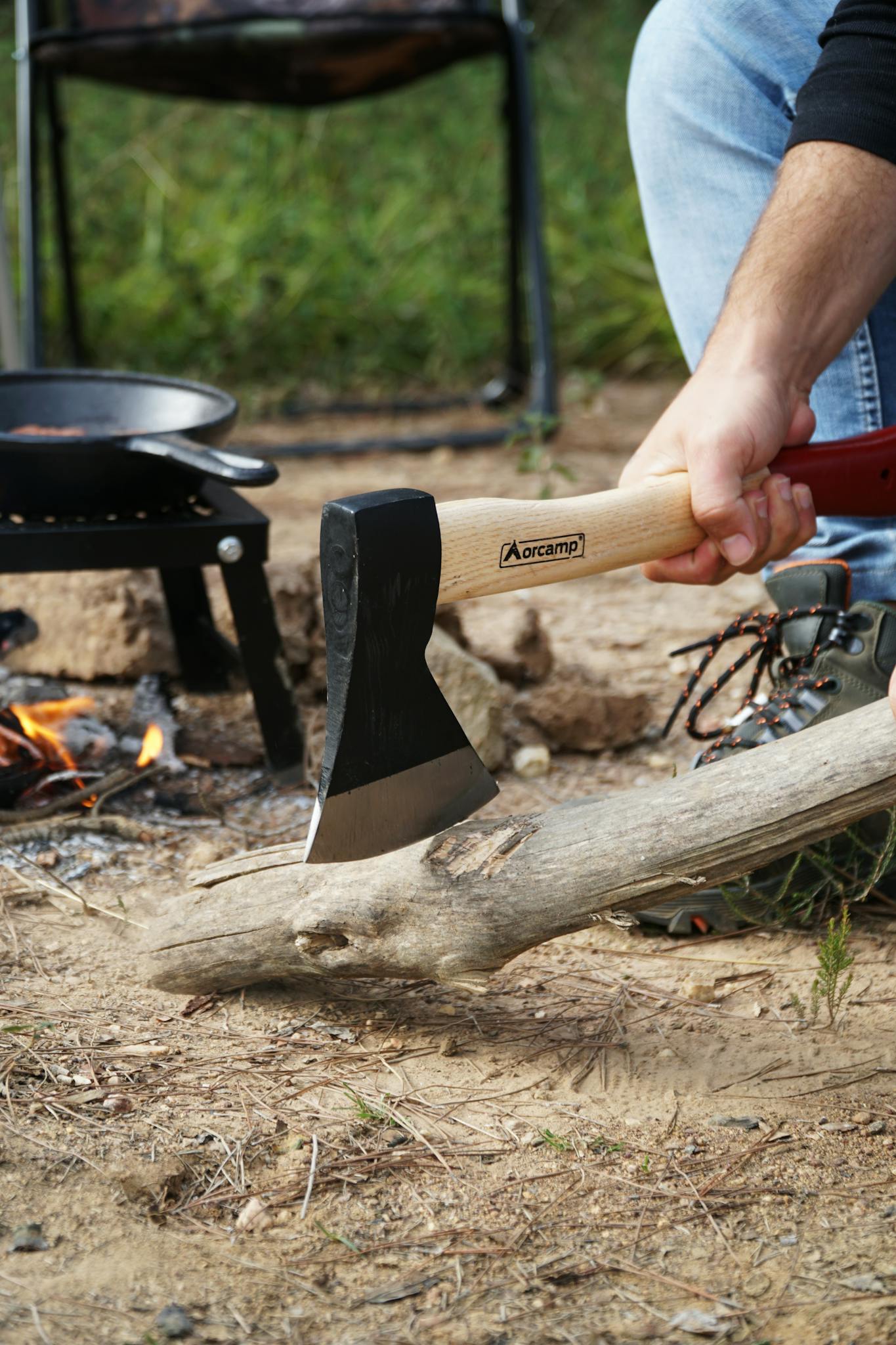Person using an axe to split wood at a campsite with a fire and cooking pan nearby.
